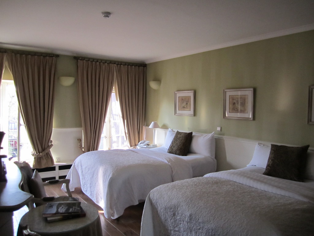 Bright guest room with twin beds, a small round table, chairs, and a clear walkway near tall windows